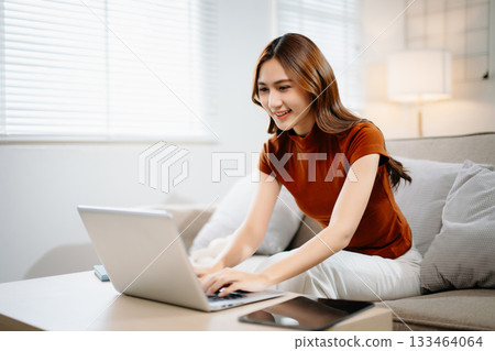 Cheerful Asian woman smiling while working on laptop at home. Modern remote work, online learning, and freelance lifestyle concept with confidence and creativity. Cheerful Asian woman smiling while working on laptop at home. Modern remote work, online learning, and freelance lifestyle concept with confidence and creativity. 133464064