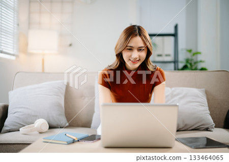 Cheerful Asian woman smiling while working on laptop at home. Modern remote work, online learning, and freelance lifestyle concept with confidence and creativity. 133464065