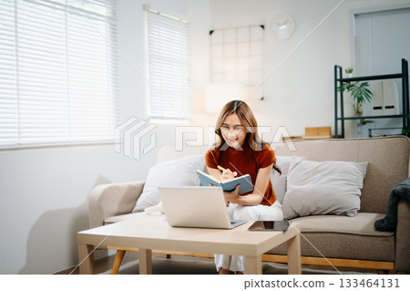 Smiling Asian woman sitting on sofa, thinking and writing notes in notebook. Creative idea, planning, education, and inspiration concept in modern home environment. 133464131