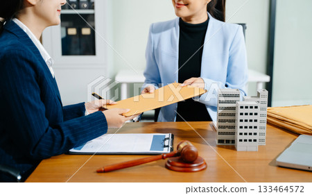Businesswoman hands over legal envelope beside building model, gavel, and contract. Real estate law, attorney advice, and document exchange in a modern office setting. Businesswoman hands over legal envelope beside building model, gavel, and contract. Real estate law, attorney advice, and document exchange in a modern office setting. 133464572