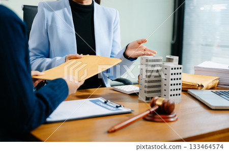 Businesswoman hands over legal envelope beside building model, gavel, and contract. Real estate law, attorney advice, and document exchange in a modern office setting. 133464574