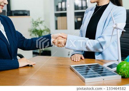Two businesswomen shake hands in an eco office with wind and solar models, symbolizing green energy, clean tech, and sustainable business success. 133464582