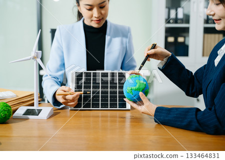 Two women analyze eco globe models, solar panels, and a smart home to plan renewable energy solutions for a greener, sustainable future. 133464831
