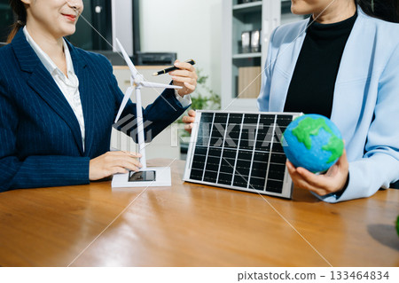 Two businesswomen shake hands in an eco office with wind and solar models, symbolizing green energy, clean tech, and sustainable business success. 133464834
