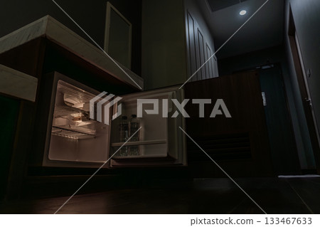 A small fridge glows softly in a dim hotel room as its open door reveals interior lighting. The low-angle view highlights the contrast between darkness and warm light. A small fridge glows softly in a dim hotel room as its open door reveals interior lighting. The low-angle view highlights the contrast between darkness and warm light. 133467633