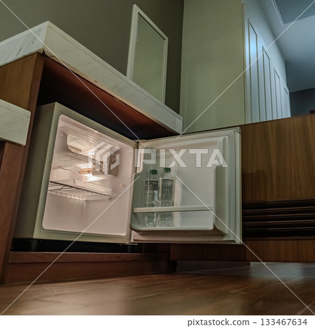 A compact refrigerator stands open in a dark hotel room with only its inner light visible. The faint glow adds a quiet atmosphere to the shadowed space. 133467634