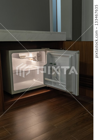 A compact refrigerator stands open in a dark hotel room with only its inner light visible. The faint glow adds a quiet atmosphere to the shadowed space. 133467635