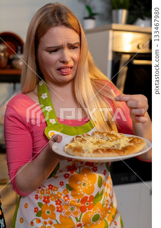 Blonde woman in apron grimaces at burned noodles in kitchen, Home cook with floral apron expressing disappointment over overcooked spaghetti in bright kitchen setting 133467980