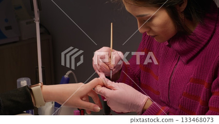 Manicure process. Polygel technique. Female beautician hands in latex gloves drawing varnish on clients nail tip with thin brush salon routine. Manicure process. Polygel technique. Female beautician hands in latex gloves drawing varnish on clients nail tip with thin brush salon routine. 133468073