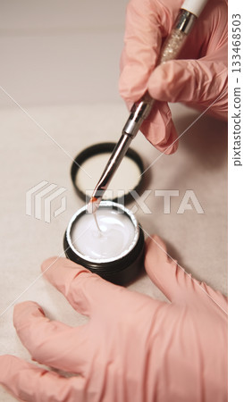 Fingernails gel. Acrylic hand manicure. Female beautician in gloves holding brush over nail polish mixing fluid getting ready for beauty procedure. Fingernails gel. Acrylic hand manicure. Female beautician in gloves holding brush over nail polish mixing fluid getting ready for beauty procedure. 133468503