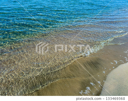 Waves gently lap the sandy shoreline under a clear blue sky on a sunny day at the beach 133469368