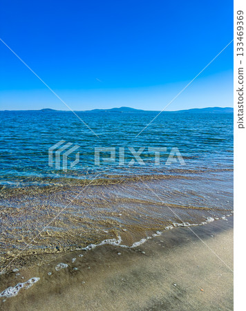 Waves gently lap against the sandy shore under a bright blue sky with distant islands on the horizon during a sunny afternoon at the beach 133469369