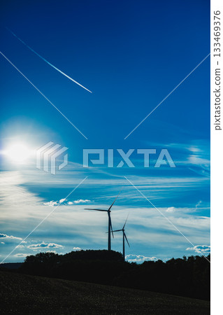 Vibrant evening sky contrasts with modern wind turbines on a serene landscape near rolling hills and a distant tree line Vibrant evening sky contrasts with modern wind turbines on a serene landscape near rolling hills and a distant tree line 133469376