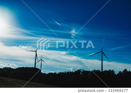 Wind turbines stand tall against a clear blue sky with wispy clouds and a bright sun illuminating the landscape in the background 133469377