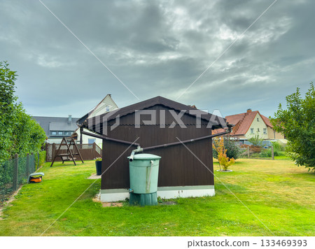 Charming garden scene featuring a brown wooden shed with a green water tank under a cloudy sky in a peaceful neighborhood Charming garden scene featuring a brown wooden shed with a green water tank under a cloudy sky in a peaceful neighborhood 133469393