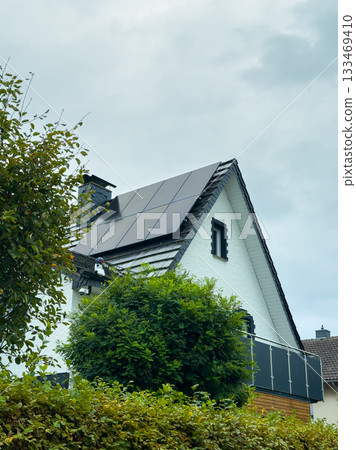 Modern home with solar panels nestled among green trees under a cloudy sky, showcasing a blend of nature and sustainable energy 133469410