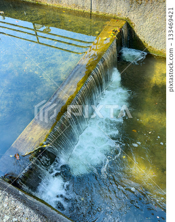 Water flows gently over a stone edge in a serene, sunlit stream surrounded by tranquil nature in a peaceful outdoor setting Water flows gently over a stone edge in a serene, sunlit stream surrounded by tranquil nature in a peaceful outdoor setting 133469421