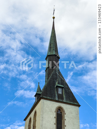 Stunning tall church steeple reaching into the blue sky against a backdrop of soft white clouds, capturing a moment of serene faith and architecture during daylight Stunning tall church steeple reaching into the blue sky against a backdrop of soft white clouds, capturing a moment of serene faith and architecture during daylight 133469423