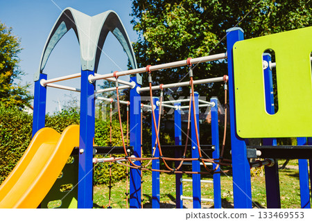 Bright and colorful playground structure with a slide, climbing ropes, and green accents under a sunny sky 133469553