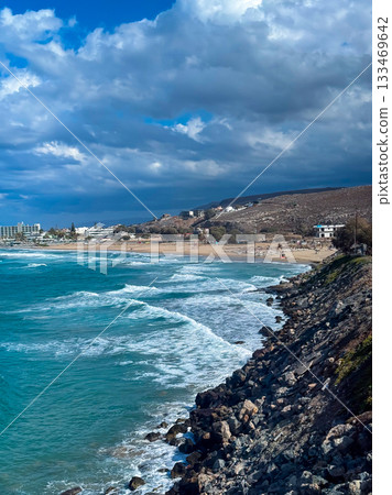 Waves crashing against rocky shoreline under a dramatic sky near a tranquil beach at sunset 133469642