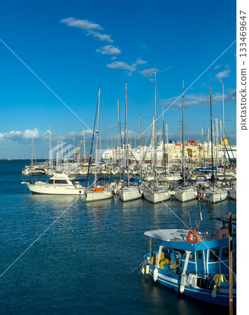 Sailboats sway gently in the sunlit harbor of a vibrant seaside town on a clear day, with clouds drifting above and city buildings in the background Sailboats sway gently in the sunlit harbor of a vibrant seaside town on a clear day, with clouds drifting above and city buildings in the background 133469647