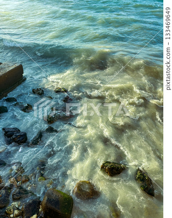 Waves gently caress the rocky shore as sunlight dances on the water's surface near a tranquil coastal landscape Waves gently caress the rocky shore as sunlight dances on the water's surface near a tranquil coastal landscape 133469649
