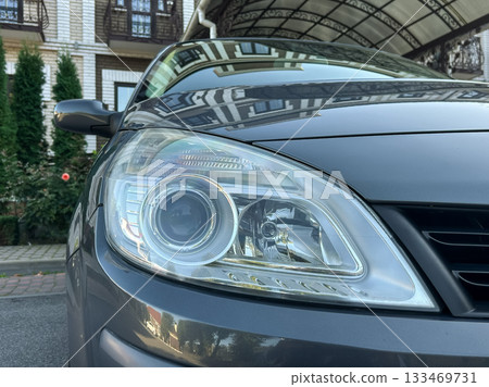 Close-up view of a modern car's headlight showcasing sleek design against a beautiful house backdrop in the afternoon light 133469731