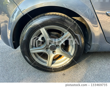 Flat tire on a silver car parked in a sunny urban street during the afternoon, showcasing the damaged wheel and tire 133469735