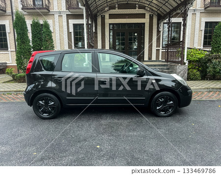 Sleek black compact car parked outside an elegant building surrounded by lush greenery on a sunny day in a peaceful neighborhood 133469786