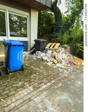 Construction debris accumulates beside blue and black waste bins in a quiet urban setting during late afternoon Construction debris accumulates beside blue and black waste bins in a quiet urban setting during late afternoon 133469794