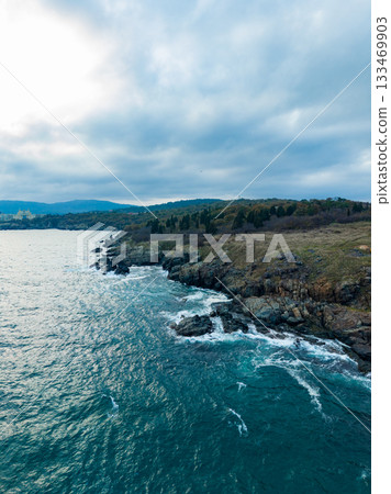Waves crashing against rocky shoreline under a cloudy sky at dusk by the coast Waves crashing against rocky shoreline under a cloudy sky at dusk by the coast 133469903