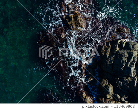 Waves crashing against rugged rocks at the shoreline during a sunny afternoon by the ocean Waves crashing against rugged rocks at the shoreline during a sunny afternoon by the ocean 133469908