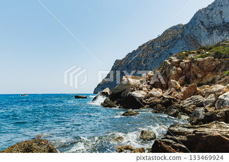 Waves crash against rocky cliffs under a clear blue sky on a sunny day by the coast, creating a serene and natural landscape 133469924