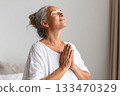 A serene, silver haired woman closes her eyes and lifts her face gently upward, fingers pressed together in gratitude, a quiet moment of wellness and hormonal balance during midlife transition A serene, silver haired woman closes her eyes and lifts her face gently upward, fingers pressed together in gratitude, a quiet moment of wellness and hormonal balance during midlife transition 133470329