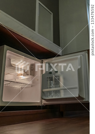 A small refrigerator sits inside a wooden cabinet with its door open, and soft interior light highlights the empty shelves and the bottle inside. 133476592