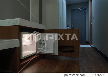 A small refrigerator sits inside a wooden cabinet with its door open, and soft interior light highlights the empty shelves and the bottle inside. 133476595