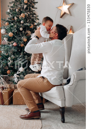 Father shares a joyful moment with his baby son in front of a decorated Christmas tree during a festive holiday celebration at home 133478830