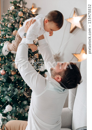 Father joyfully lifts his young child in front of a decorated Christmas tree with warm lights and festive ornaments during the holiday season 133478832