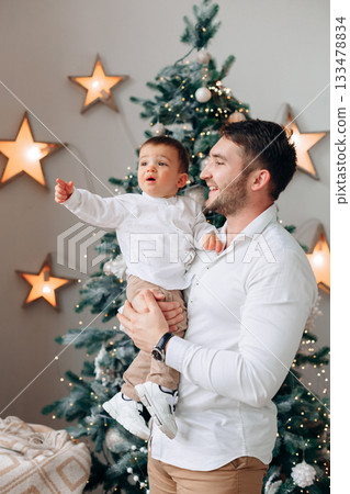 Father holds his joyful son in front of a decorated Christmas tree filled with lights and stars while celebrating the holiday season together Father holds his joyful son in front of a decorated Christmas tree filled with lights and stars while celebrating the holiday season together 133478834