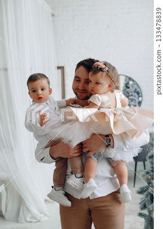 Father holding his children in a cozy indoor setting during a festive celebration in the morning light Father holding his children in a cozy indoor setting during a festive celebration in the morning light 133478839