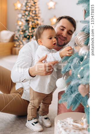 Father and child joyfully decorate a Christmas tree together in a cozy living room filled with festive lights and decorations 133478855