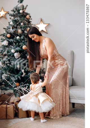 Mother in sparkling dress helps little girl near decorated Christmas tree with gifts during festive season 133478867