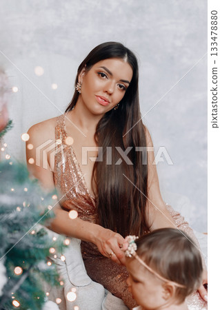 Beautiful woman in elegant dress sits beside child during festive celebration in cozy indoor setting with soft lighting and holiday decorations 133478880