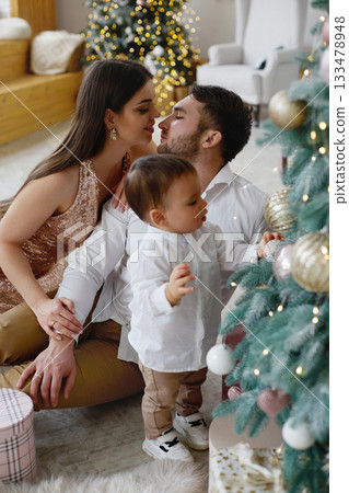 Couple and child together celebrating Christmas in a cozy living room with decorations and a beautifully adorned tree Couple and child together celebrating Christmas in a cozy living room with decorations and a beautifully adorned tree 133478948