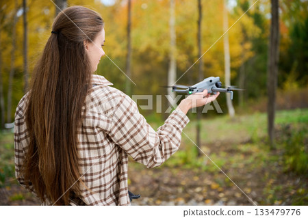 A young woman stands in a colorful autumn forest, preparing to launch her drone. Surrounding trees display bright yellow and orange leaves, creating a stunning backdrop. 133479776