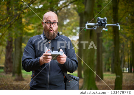 In a colorful forest filled with autumn leaves, a man excitedly operates a drone with a remote. His surprise showcases the thrill of flying technology in nature. 133479778