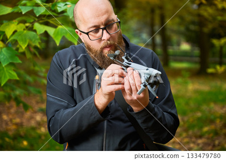 A person intently inspects a small drone while surrounded by vibrant autumn trees. Sunlight filters through the leaves, creating a peaceful atmosphere. 133479780