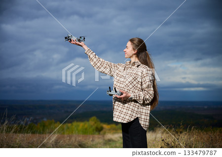 A young woman stands on a grassy hill, holding a drone in one hand and its controller in the other. She's ready for a thrilling flight against a stunning backdrop of cloudy skies. 133479787