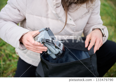 A woman carefully places a compact drone into a black bag while in a scenic outdoor setting. She is ready for an exciting day of capturing aerial views. A woman carefully places a compact drone into a black bag while in a scenic outdoor setting. She is ready for an exciting day of capturing aerial views. 133479790