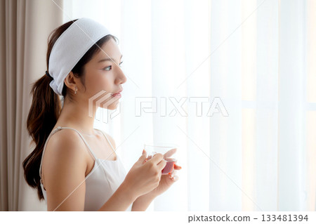 Beautiful young asian woman with headband enjoying tea standing near window with satisfied. 133481394